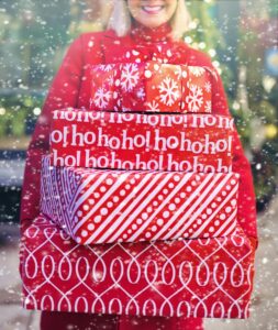 Une femme tenant joyeusement une pile de cadeaux de Noël rouges dans un décor extérieur enneigé.